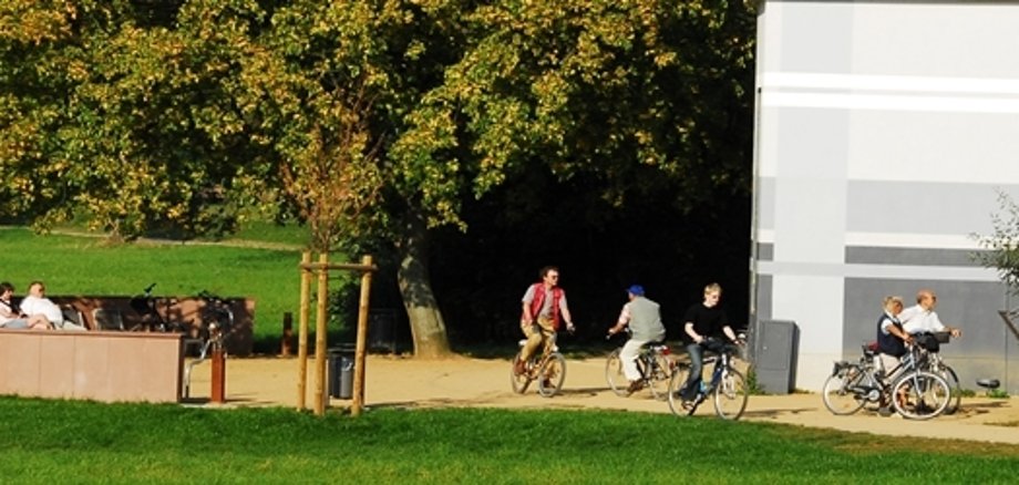 Gruppe von Radfahrern auf einem Weg neben einer Wiese und einem weißen Gebäude im Hintergrund
