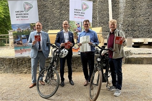 Vier Männer in Freizeitkleidung stehen mit Fahrrädern vor zwei Bannern mit der Aufschrift 'Kneipp Bäder Eck Wetterau' und halten Karten oder Broschüren in der Hand