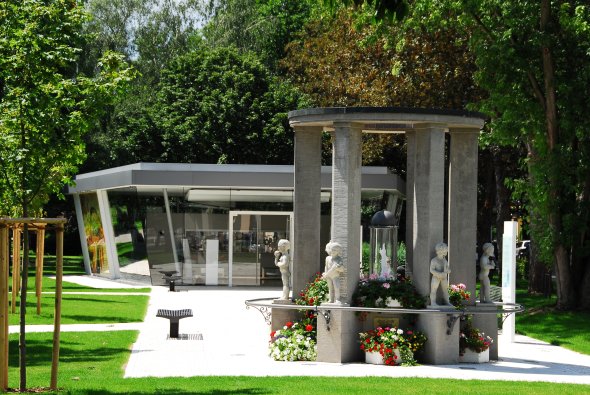 Brunnen mit römischen Figuren und Blumen vor modernem Glasgebäude in Parkanlage