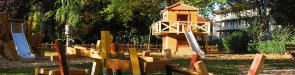 Spielplatz mit mehreren Holzspielgeräten, darunter Rutschen und Wippen, umgeben von Bäumen und Wohngebäuden