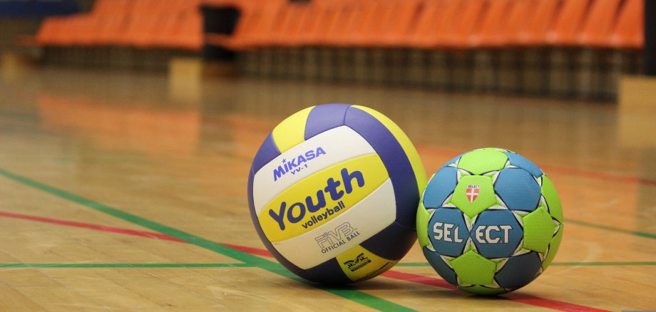 Zwei Volleyballbälle auf dem Boden einer Sporthalle vor leeren orangenen Tribünen