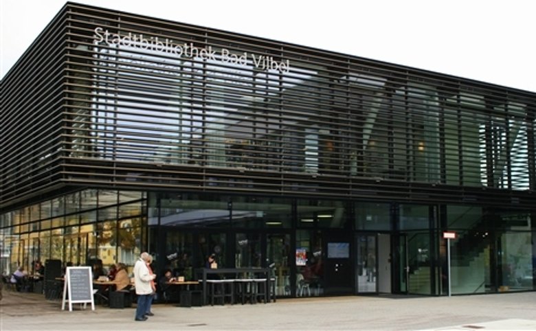 Modernes Gebäude mit Glasfassade und horizontalen Lamellen, davor Menschen und Sitzgelegenheiten