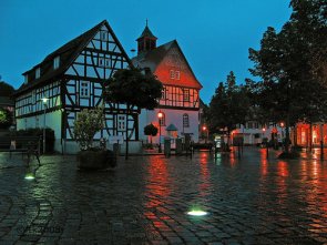 Fachwerkhaus an einem gepflasterten Platz mit Bänken und Bäumen bei Dämmerung mit beleuchteten Bodenelementen