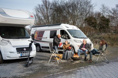 Vier Personen sitzen auf Campingstühlen vor zwei Wohnmobilen um ein kleines Lagerfeuer auf einem Stellplatz