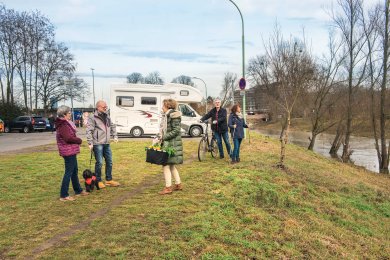 Fünf Personen stehen auf einem grasbewachsenen Weg neben einem Wohnmobil und einem Fluss, eine Person hält ein Fahrrad