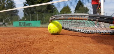 Tennisball liegt auf rotem Sandplatz neben Tennisracket, im Hintergrund Netz und Spieler in rotem Oberteil