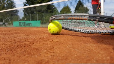 Tennisball liegt auf rotem Sandplatz neben Tennisracket, im Hintergrund Netz und Spieler in rotem Oberteil