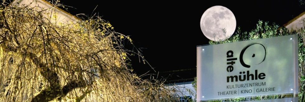Beleuchteter Baum vor einem Schild mit der Aufschrift 'Alte Mühle Kulturzentrum Theater Kino Galerie' und Vollmond am Nachthimmel