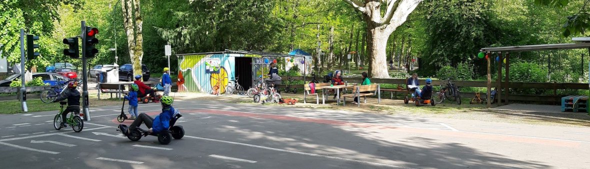 Kinder mit Helmen fahren auf Dreirädern und Fahrrädern auf einem Verkehrsspielplatz mit Straßenmarkierungen und Sitzbänken unter Bäumen
