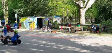 Verkehrsspielplatz Kinder mit Helmen fahren auf Dreirädern und Fahrrädern auf einem Verkehrsspielplatz mit Straßenmarkierungen und Sitzbänken unter Bäumen