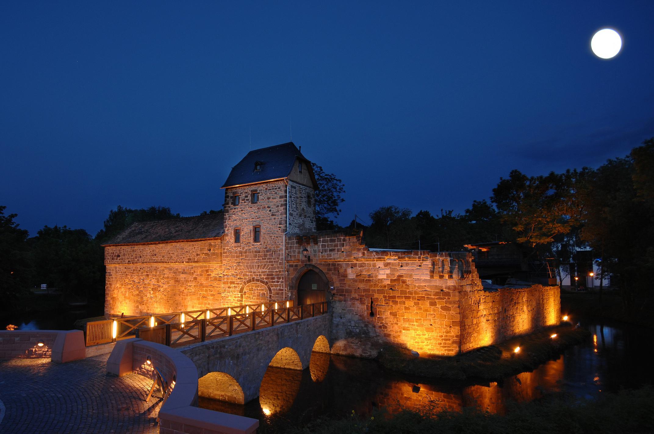Beleuchtete mittelalterliche Wasserburg bei Nacht unter klarem Himmel mit sichtbarem Vollmond