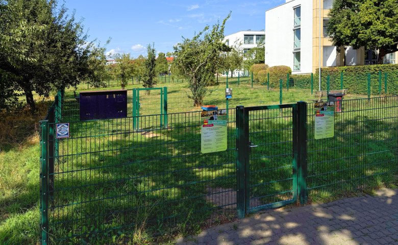 Eingezäunte Hundewiese mit grüner Wiese, Bäumen und Informationsschildern vor einem modernen Gebäude