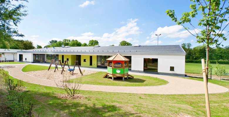 Flacher moderner Kindergartenbau mit Spielgeräten und kleinem Pavillon auf einem weitläufigen Außengelände unter blauem Himmel