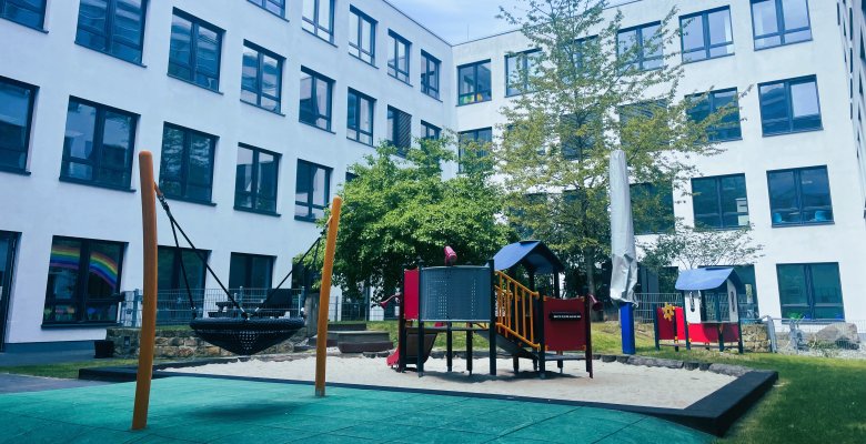 Spielplatz eines Kindergartens mit Sandkasten, Klettergerüst und Nestschaukel vor einem mehrstöckigen weißen Gebäude mit vielen Fenstern