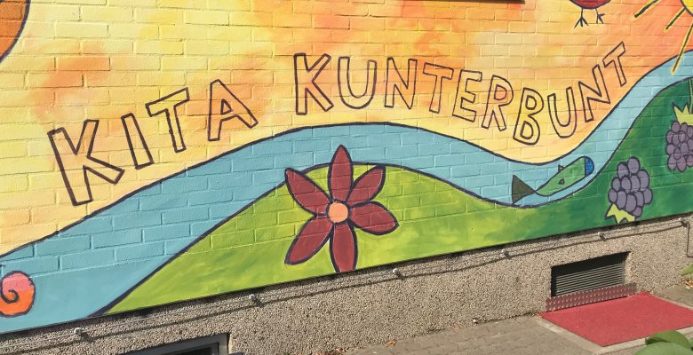 Bunte Wandbemalung mit Schriftzug 'KITA KUNTERBUNT', Sonne, Vogel, Fluss, Blume und Trauben an einer Hauswand