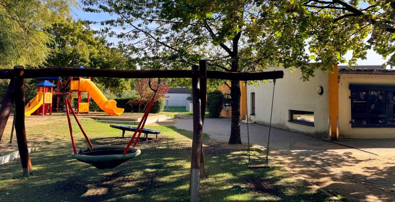 Spielplatz mit Nestschaukel, einfacher Schaukel, Klettergerüst mit Rutsche und Bäumen neben einem hellen Kita-Gebäude