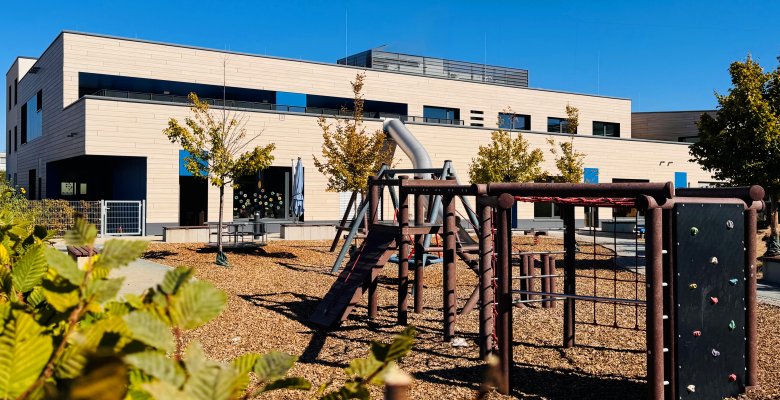 Moderner zweigeschossiger Gebäudekomplex mit hellem Fassadenmaterial und großem Außenspielplatz mit Klettergerüst und Rutsche im Vordergrund