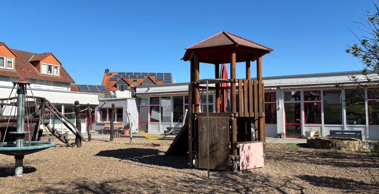 Spielplatz mit Kletterturm und Nestschaukel vor einem eingeschossigen Gebäude unter blauem Himmel