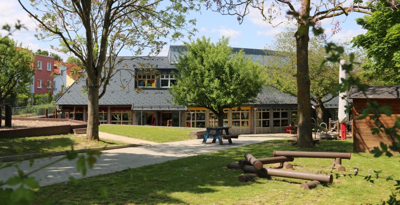 Gebäude einer Kindertagesstätte mit großem Garten, Bäumen, Sitzbank und Spielgeräten auf einer Wiese