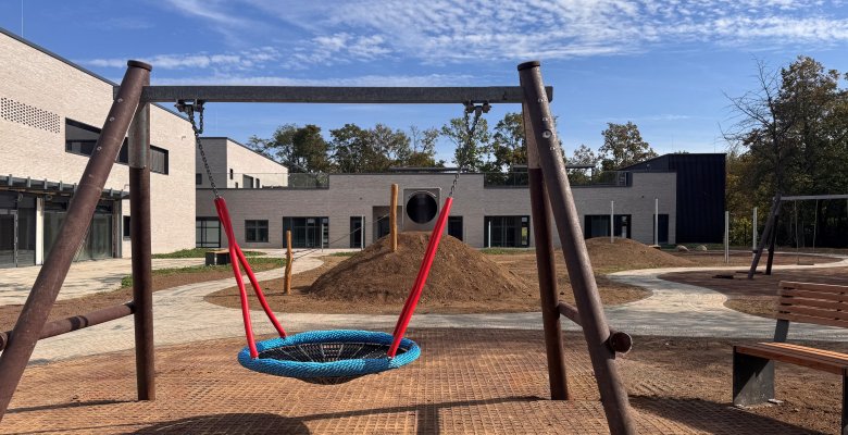 Nestschaukel mit roten Seilen auf einem Spielplatz vor einem modernen eingeschossigen Kita-Gebäude unter blauem Himmel