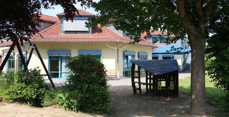 Gelbes Gebäude mit rotem Ziegeldach und blau umrahmten Fenstern, davor ein Spielplatz mit Holzhaus und Schaukel unter Bäumen