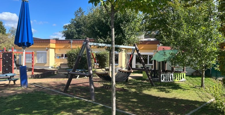 Spielplatz mit Schaukel, Klettergerüst, Sonnenschirm und einem kleinen Spielhaus vor einem eingeschossigen Kita-Gebäude unter blauem Himmel