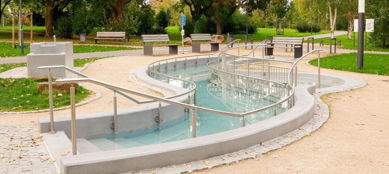 Park mit geschwungenem Wasserbecken, umgeben von Bänken und großen Bäumen mit herbstlichen Blättern