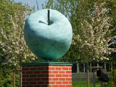 Große Apfel-Skulptur auf einem quadratischen Sockel aus roten Ziegelsteinen vor blühenden Bäumen