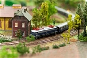 Foto: Modellbahn mit blauer Lokomotive und mehreren Waggons auf Gleisen vor einem braunen Haus mit Schornstein.