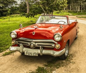 Roter Oldtimer-Cabrio steht auf unbefestigter Straße vor grünem Wald unter bewölktem Himmel