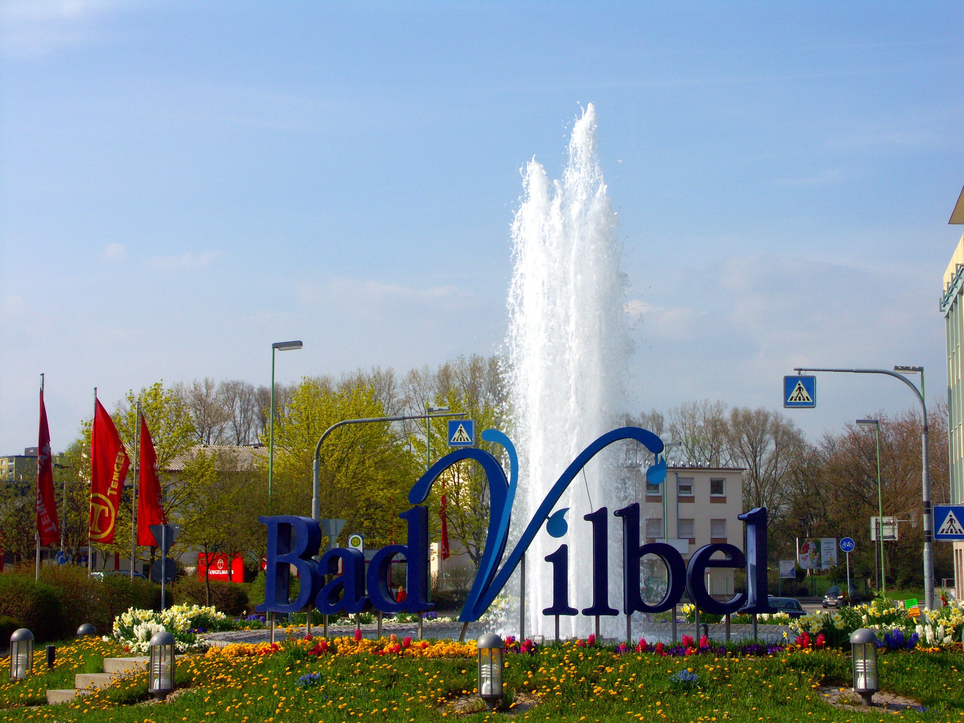 Springbrunnen mit blauem Schriftzug 'Bad Vilbel' umgeben von Blumen und roten Fahnen im Hintergrund