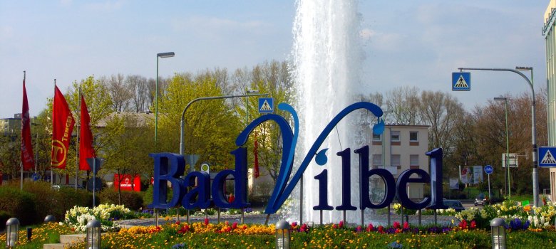 Günther-Biwer-Kreisel Springbrunnen mit blauem Schriftzug 'Bad Vilbel' umgeben von Blumen und roten Fahnen im Hintergrund