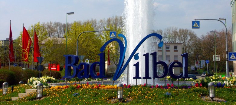 Günther-Biwer-Kreisel Springbrunnen mit blauem Schriftzug 'Bad Vilbel' umgeben von Blumen und roten Fahnen im Hintergrund