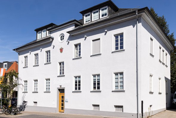 Weißes mehrstöckiges Gebäude mit Satteldach und mehreren Fenstern an einer Straßenecke