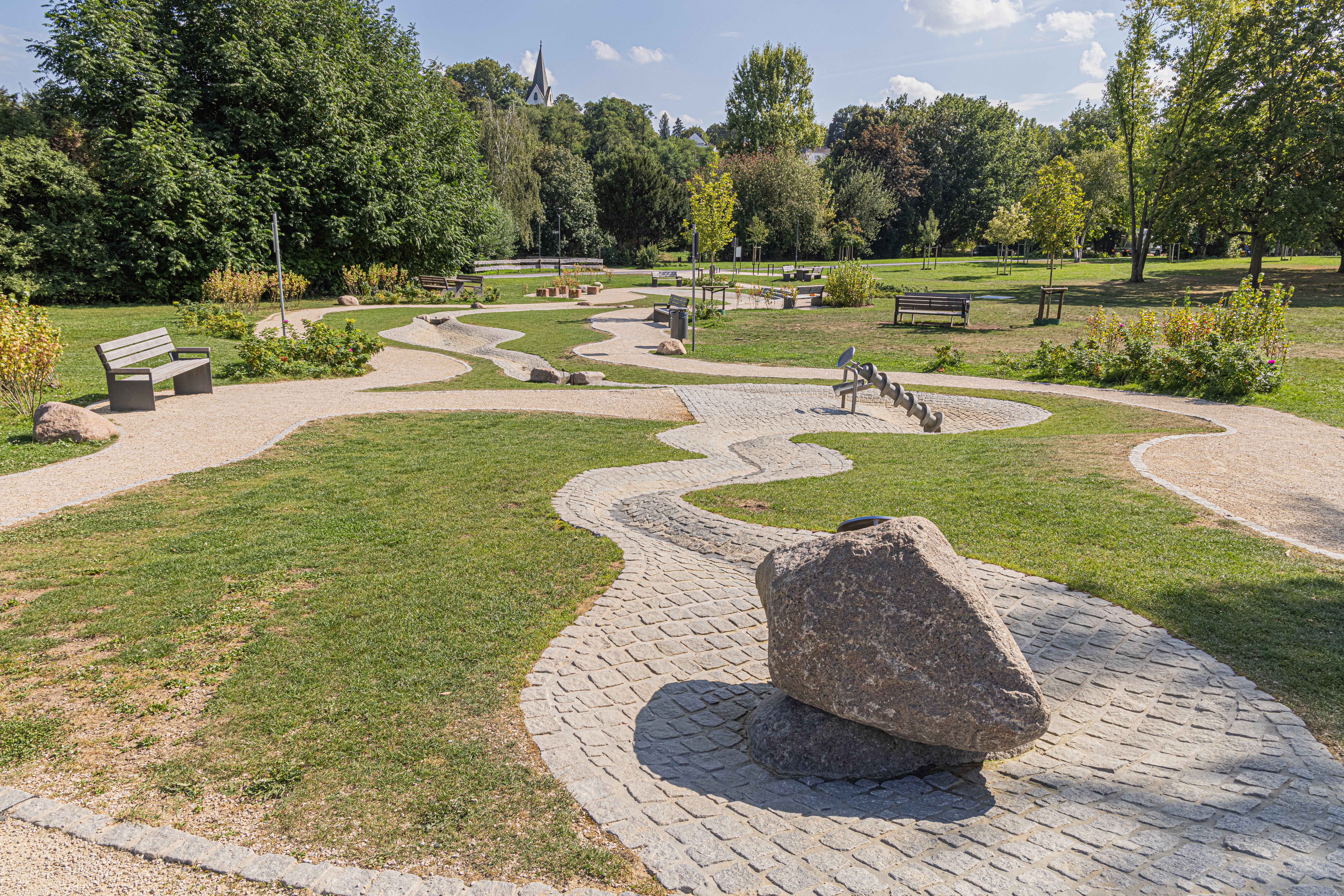 Park mit geschwungenen gepflasterten Wegen, mehreren Bänken und einem großen Stein auf einem gepflasterten Kreis