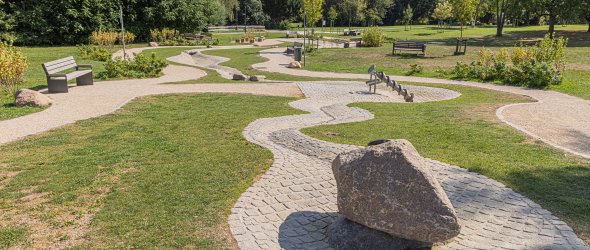 Park mit geschwungenen gepflasterten Wegen, mehreren Bänken und einem großen Stein auf einem gepflasterten Kreis
