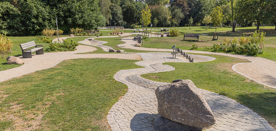 Park mit geschwungenen gepflasterten Wegen, mehreren Bänken und einem großen Stein auf einem gepflasterten Kreis