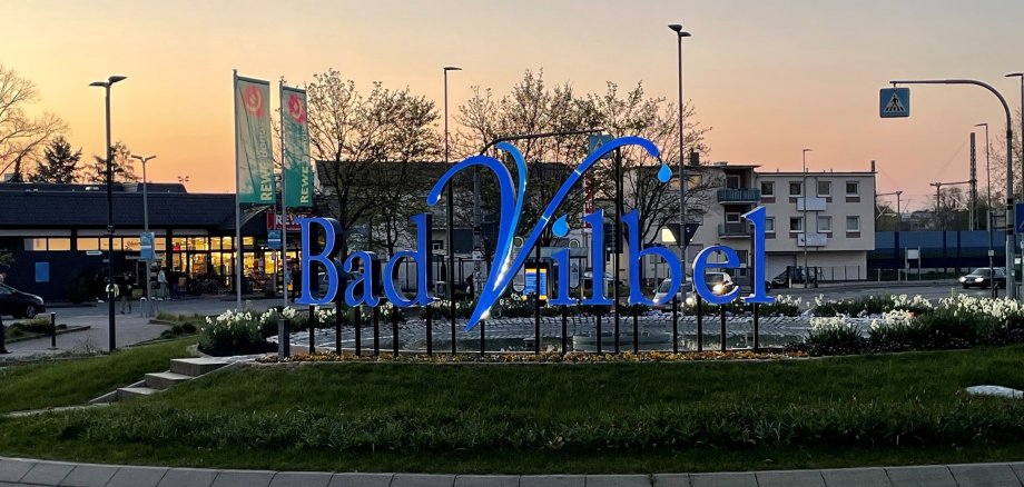 Großes blaues Schriftzug-Logo "Bad Vilbel" auf einem begrünten Kreisverkehr bei Dämmerung