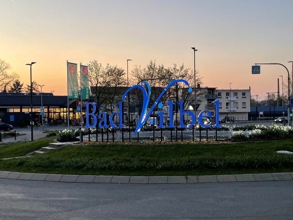 Großes blaues Schriftzug-Logo "Bad Vilbel" auf einem begrünten Kreisverkehr bei Dämmerung