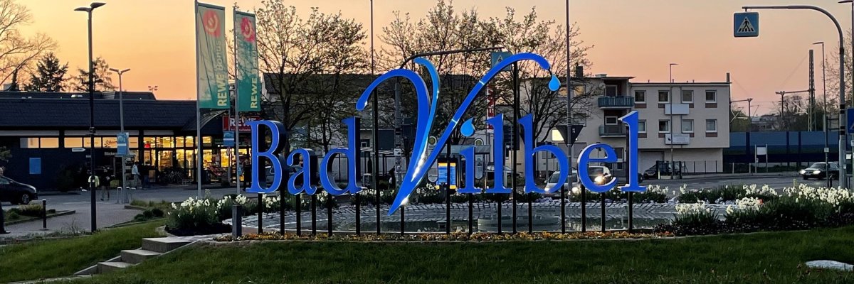 Großes blaues Schriftzug-Logo "Bad Vilbel" auf einem begrünten Kreisverkehr bei Dämmerung