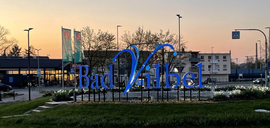 Biwerkreisel Großes blaues Schriftzug-Logo "Bad Vilbel" auf einem begrünten Kreisverkehr bei Dämmerung