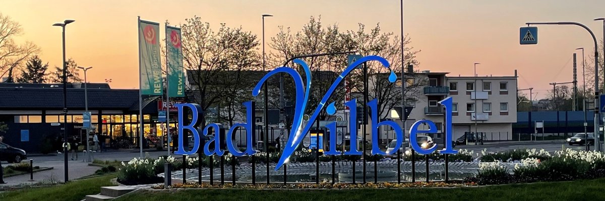 Biwerkreisel Großes blaues Schriftzug-Logo "Bad Vilbel" auf einem begrünten Kreisverkehr bei Dämmerung