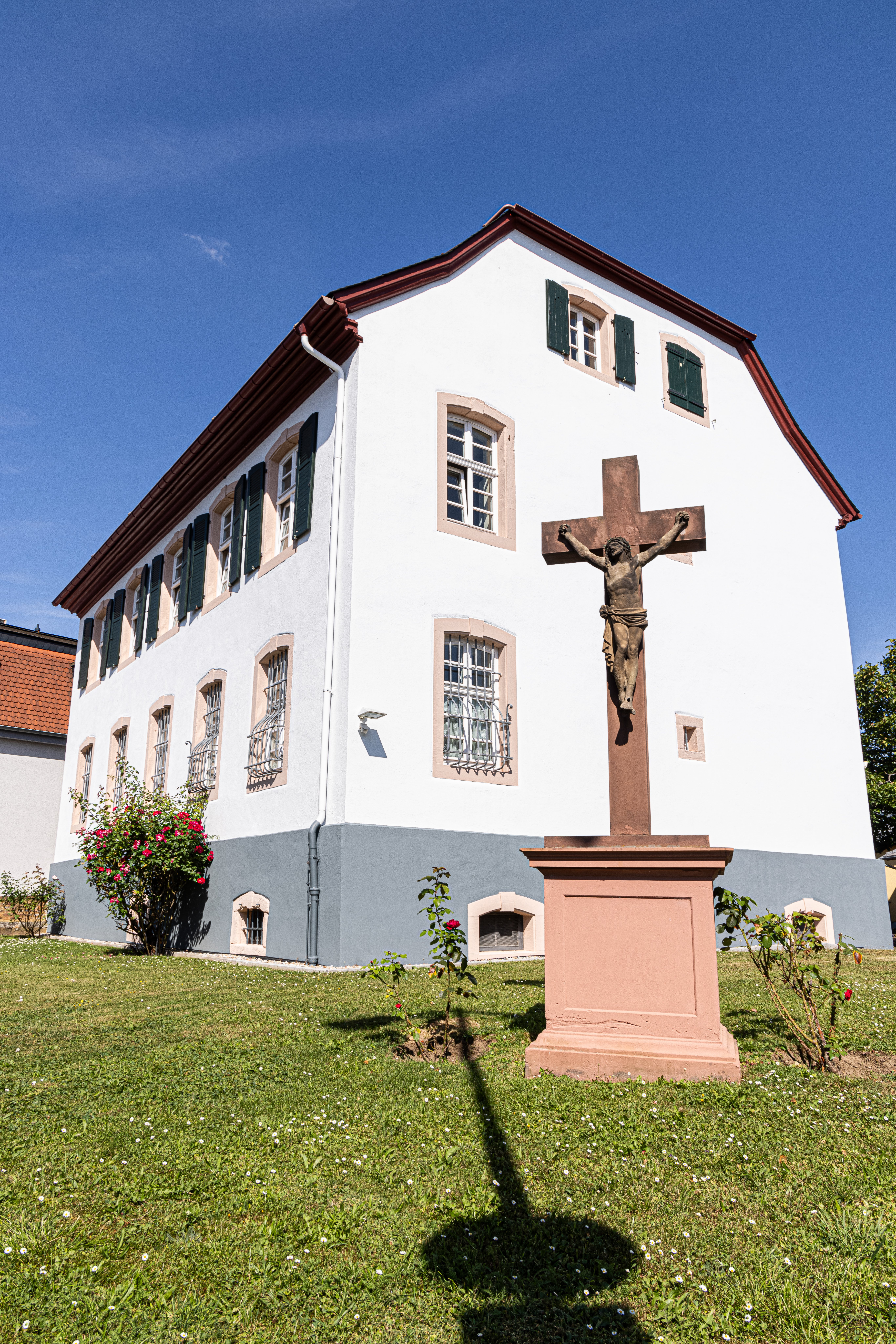 Pfarrei Bad Vilbel St. Nikolaus Weißes zweistöckiges Gebäude mit grünen Fensterläden und einem steinernen Kreuz mit Jesusfigur im Vordergrund auf einer Rasenfläche
