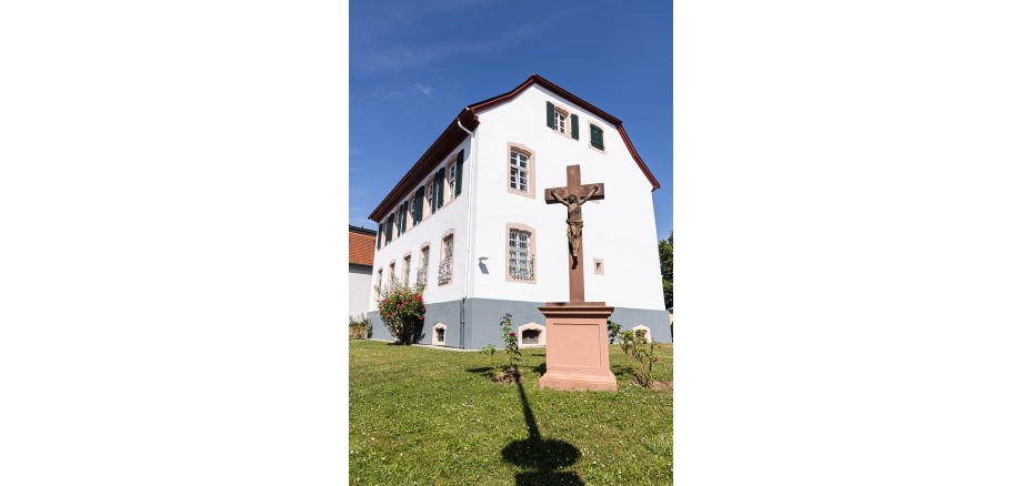 Weißes zweistöckiges Gebäude mit grünen Fensterläden und einem steinernen Kreuz mit Jesusfigur im Vordergrund auf einer Rasenfläche
