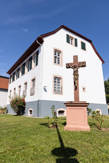 Weißes zweistöckiges Gebäude mit grünen Fensterläden und einem steinernen Kreuz mit Jesusfigur im Vordergrund auf einer Rasenfläche