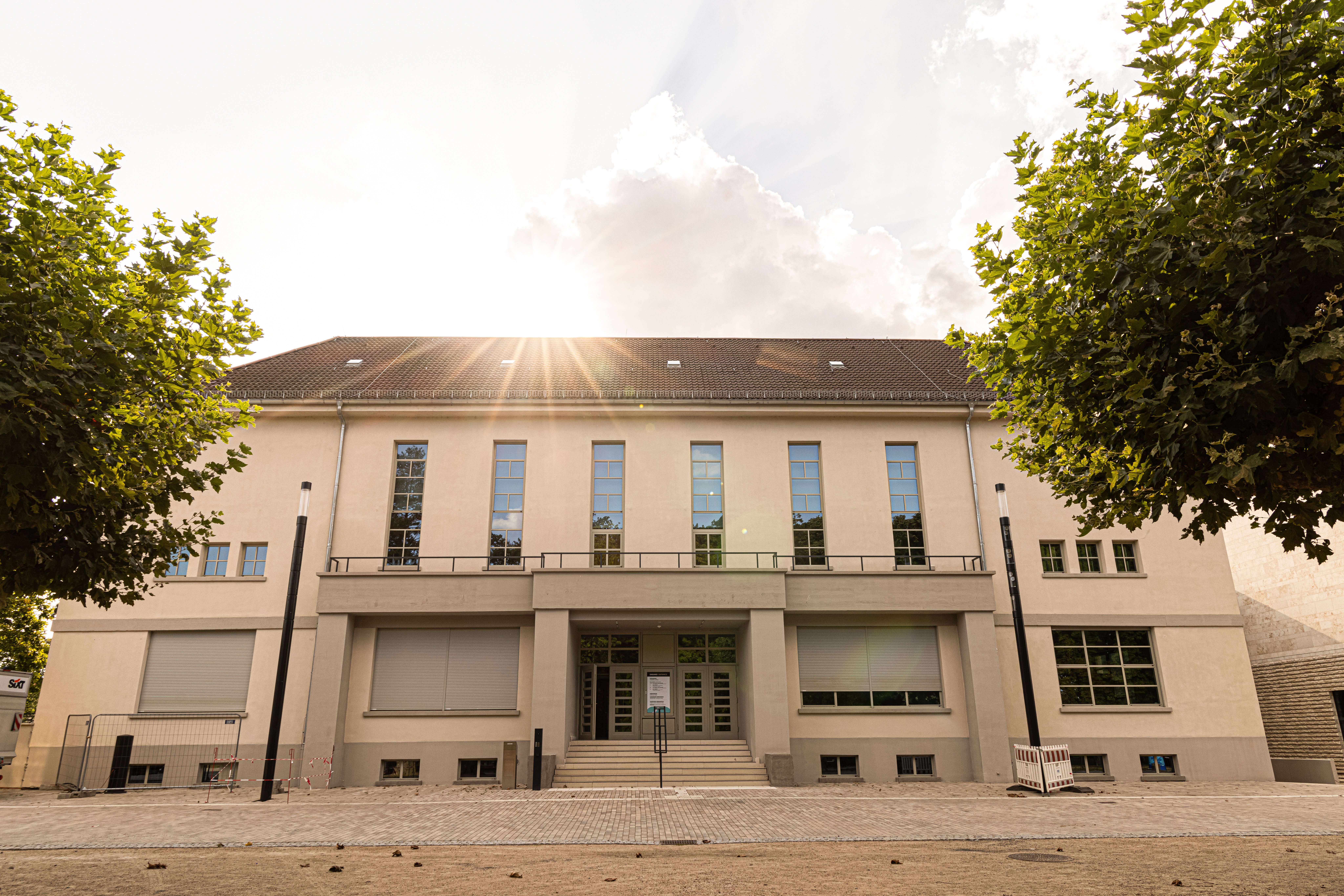 Zweistöckiges Gebäude mit mehreren Fenstern und geschlossenen Rollläden bei Sonnenlicht zwischen zwei Bäumen