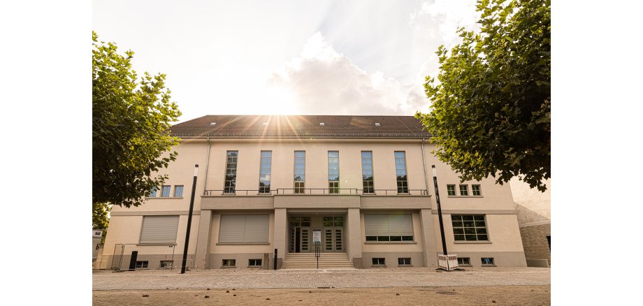 Zweistöckiges Gebäude mit mehreren Fenstern und geschlossenen Rollläden bei Sonnenlicht zwischen zwei Bäumen