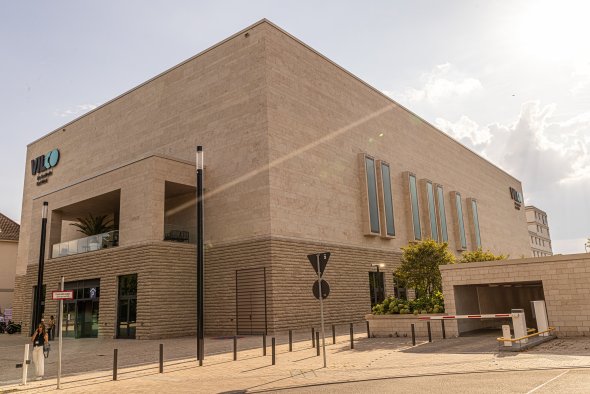 Kubisches, helles Gebäude mit vertikalen Fenstern und Logo an der Ecke an einer Straßenkreuzung bei Tageslicht