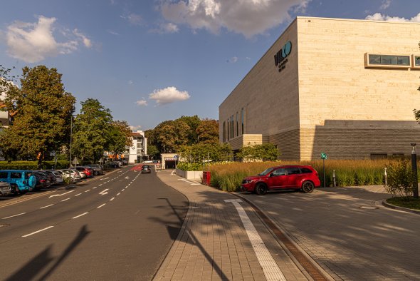 Straße mit parkenden Autos neben einem modernen Gebäude mit hellen und dunklen Fassadenflächen unter blauem Himmel mit Wolken