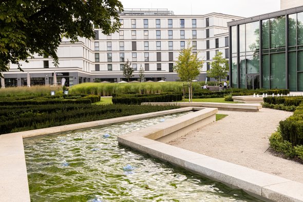Wasserbecken mit rechteckigem Verlauf vor einem modernen mehrstöckigen Gebäude und einem Glasbau mit Bäumen und Hecken im Vordergrund